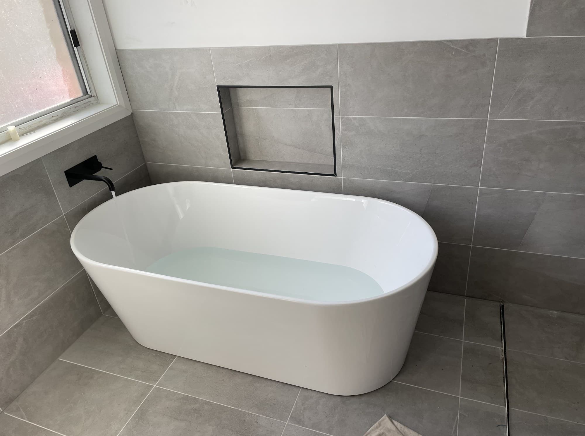 Freestanding bath plumbed in during a bathroom renovation, with matte black tapware and floor-to-ceiling tiling
